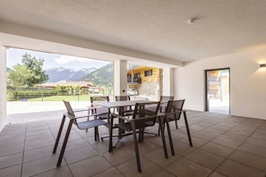 Uma sala de jantar com mesa e cadeiras, vista para montanhas e um edifício com fachada de pedra.