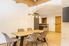 Uma zona de refeições moderna com mesa e cadeiras de madeira, um lustre pendurado e uma cozinha ao fundo.