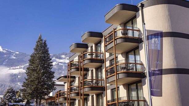 Hotel de diversos pisos amb balcons, un cartell d'estació d'esquí i un paisatge de muntanyes al fons.