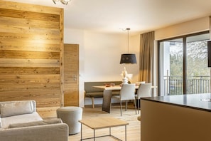 A modern interior with a wooden accent wall, a dining area with a table and chairs, a sofa, a lamp, and a view through large windows. - Apartment with 1 bedroom (St. Martin am Tennengebirge)