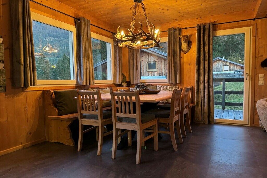 Een houten interieur met een eettafel, stoelen, een kroonluchter en een uitzicht op een blokhut door grote ramen.