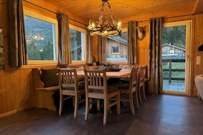 A wooden interior with a dining table, chairs, a chandelier, and a view of a cabin through large windows. - Cabin in Annaberg with swimming pool (Annaberg)