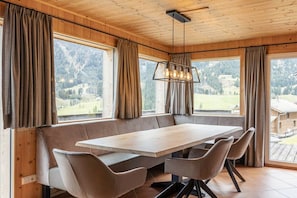 Een houten interieur met een grote tafel en stoelen, een hanglamp en grote ramen met gordijnen.