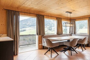 Een eetgedeelte met een houten plafond, grote ramen voorzien van gordijnen, een tafel met stoelen en een hanglamp.