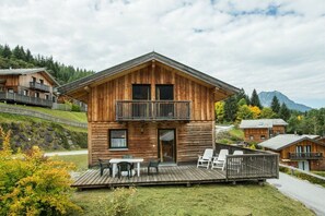 Eine Holzhütte mit Balkon und einer Terrasse mit Stühlen, vor einer Bergkulisse.