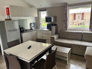 A compact kitchenette with a refrigerator, stove, microwave, and a small TV. There is a dining table with chairs and a sofa in the living area. - 5-Star Mobile Home with Shared Pools, Terrace & Private Garden in Saint-Georges-d'Oléron (Saint-Georges-d'Oléron)