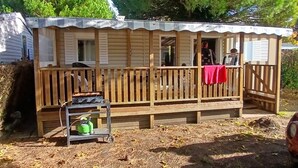 Een houten hutje met een veranda, een tafel met een brandstoftank, en een overdekte plek met een rode doek.