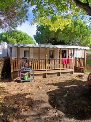 A wooden cabin with a porch, a table with a gas can, and a covered area with a red cloth. - 5-Star Mobile Home with Shared Pools, Terrace & Private Garden in Saint-Georges-d'Oléron (Saint-Georges-d'Oléron)