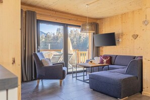 Una sala de estar moderna con un sofá, un sillón y una mesa. Hay un televisor montado en la pared y una ventana con cortinas.