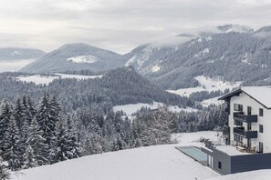 Un edificio moderno con piscina, circondato da montagne e alberi innevati.