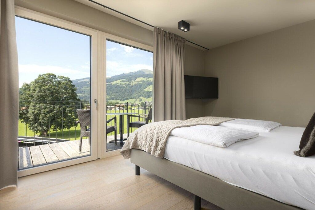 Een slaapkamer met een groot bed, een aan de muur bevestigde televisie en een glazen schuifdeur die leidt naar een balkon met uitzicht op de omgeving.