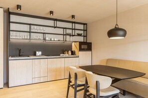 Une cuisine moderne équipée d’un four à micro-ondes encastré, d’une machine à café et d’une étagère où sont rangés des verres et des tasses. On y trouve également une table à manger avec des chaises et une suspension lumineuse au-dessus.