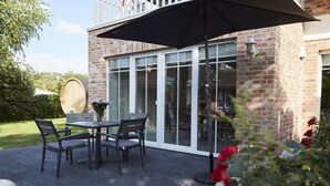 An outdoor patio with a table and chairs, a large umbrella, and a brick building with glass doors. - Holiday apartment in Büsum with swimming pool (Büsum)