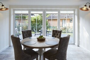 Een eethoek met een ronde tafel en vier stoelen, een schaal met fruit, en uitzicht op een tuin en gebouwen.