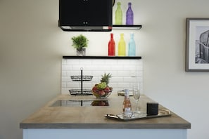 Een keuken met een houten aanrecht, een fruitschaal, een wijnglas en kleurrijke flessen op planken.