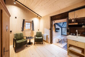A cozy room with wooden walls and a large window. There are two chairs, a small table, and a bathroom with a sink and mirror. - historic Chalet Raneburg (Matrei in Osttirol)