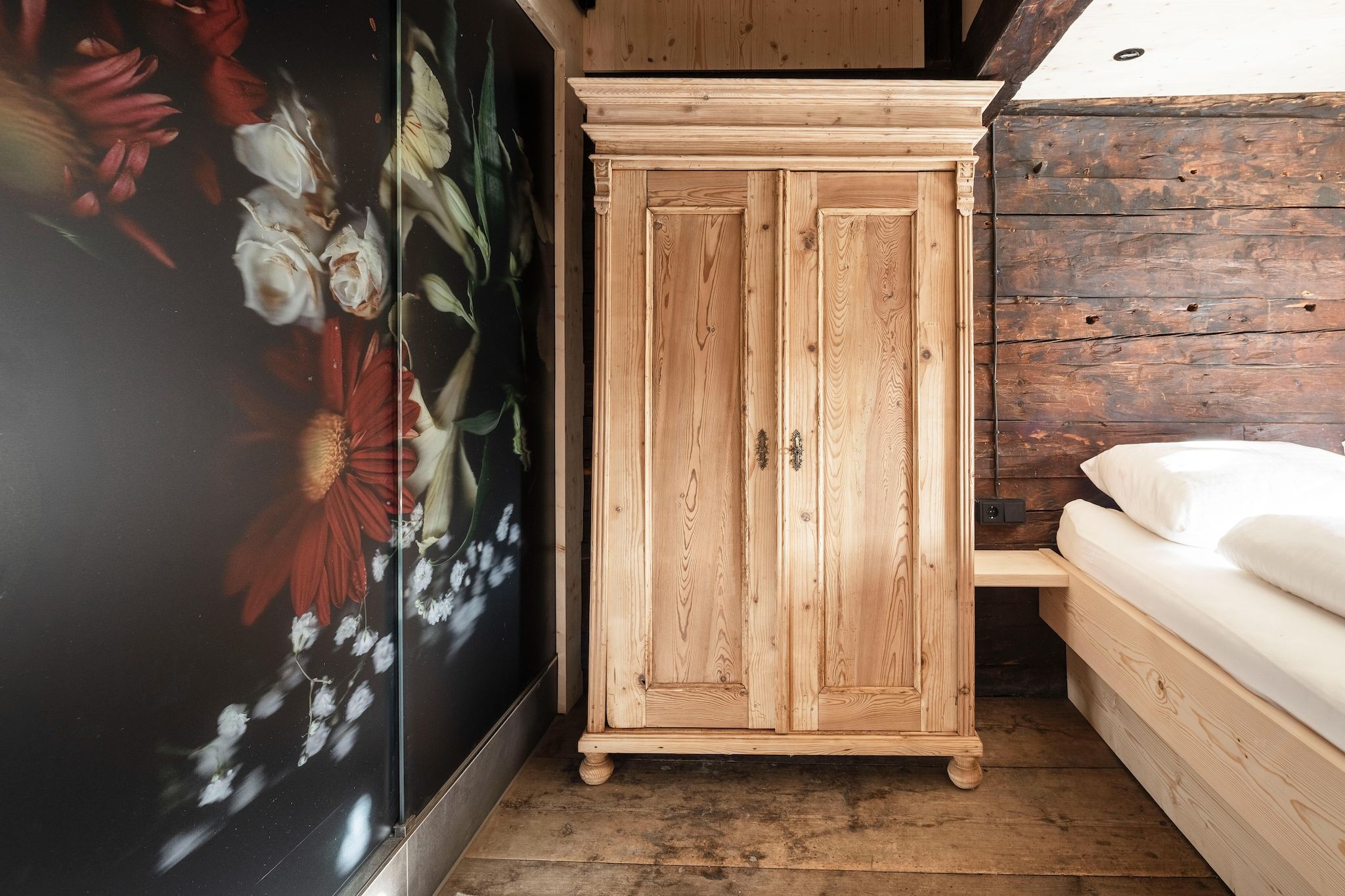 A wooden wardrobe in a room with a floral wallpaper and a bed with white bedding.