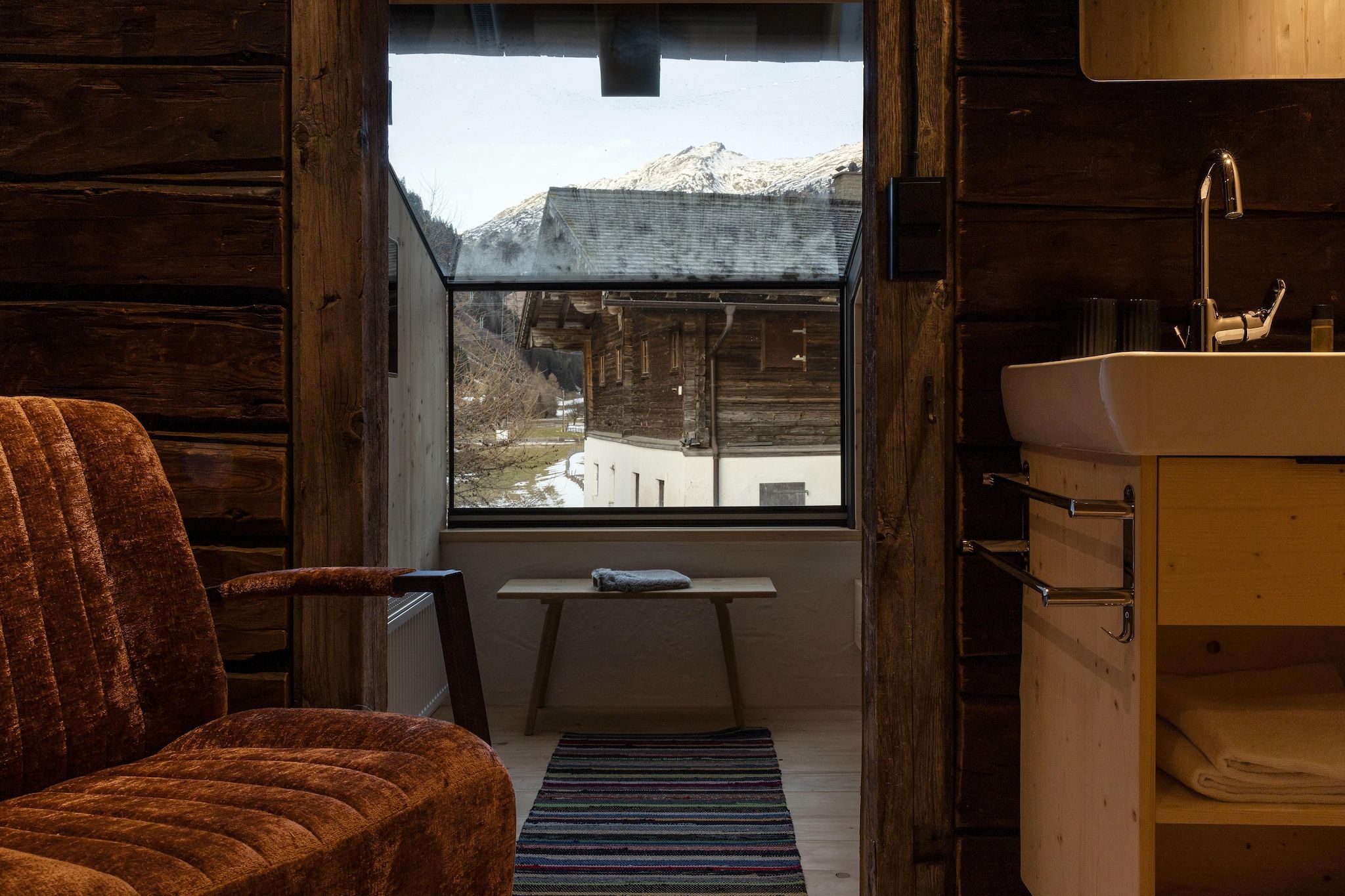 Interno rustico di una baita con una sedia di legno, un piccolo tavolo con un asciugamano, una finestra che si affaccia su un paesaggio innevato e un lavandino in legno.