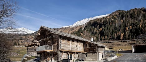 Un edificio rústico de madera con un balcón, con montañas nevadas y una ladera boscosa al fondo.