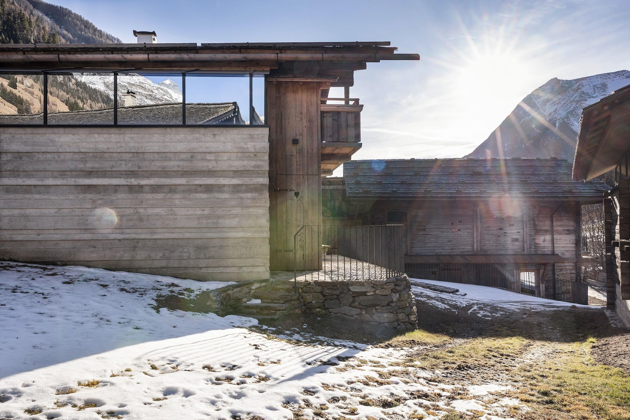 Un bâtiment en bois, doté d’un mur en pierre et d’une rambarde en métal, se dressant sur un paysage enneigé avec des montagnes en arrière-plan.