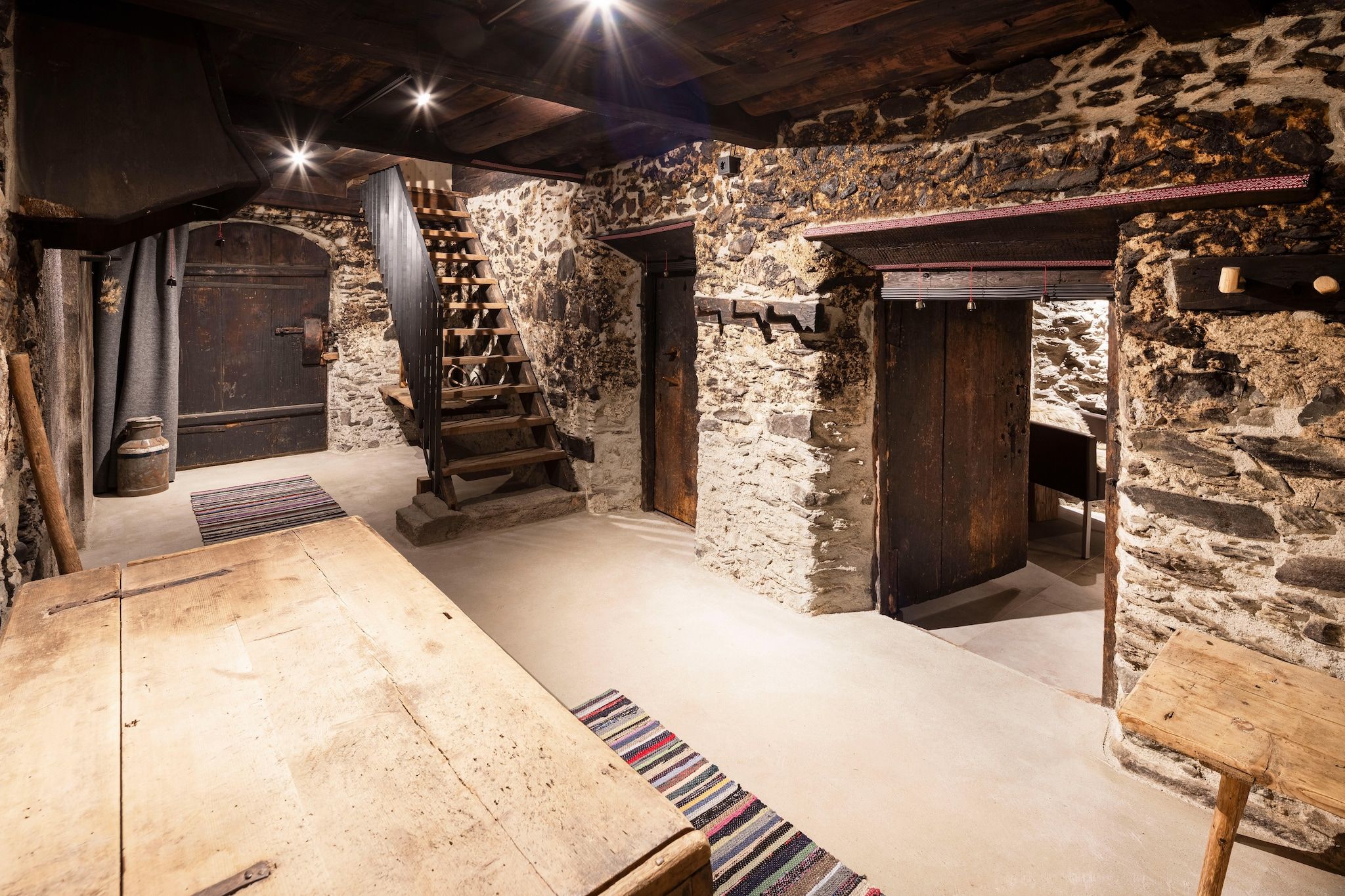 Ein rustikales Steinzimmer mit einer Holztreppe, einem langen Tisch und einer Tür.