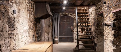 Una bodega rústica con paredes de piedra, un banco de madera, una puerta metálica y una escalera de madera que conduce a un nivel superior.