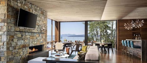 A modern living room with a stone fireplace, a flat-screen TV, and a view of the sea.
