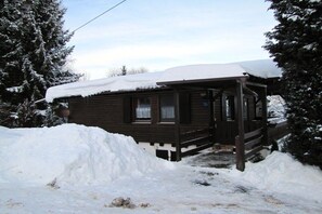 A wooden cabin with a covered porch, surrounded by snow-covered trees.