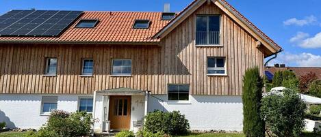 A two-story wooden house with a sloped roof, solar panels, and a small front porch.