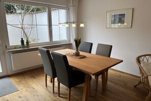 A dining room with a wooden table, four chairs, a vase with flowers, a picture on the wall, and a window with a view of trees. - Holiday apartment in Stetten with barbecue (Stetten)