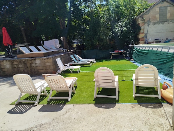 A backyard with artificial grass, white lounge chairs, and a pool area.