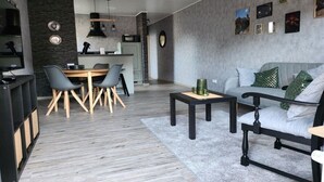 Moderni olohuone, jossa on harmaa sohva, ruokapöytä tuoleineen ja taustalla keittiötila.