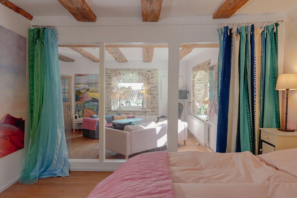 A bedroom with a bed, a window with curtains, and a view of an adjacent room with a sofa and a painting.