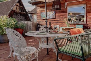 Eine Terrasse im Freien mit einem runden Tisch, weißen verzierten Stühlen und einem Korbstuhl.