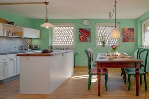 Eine Küche mit einer zentralen Kochinsel, einem Esstisch mit Stühlen und einem Fenster mit einem gemusterten Rollo.