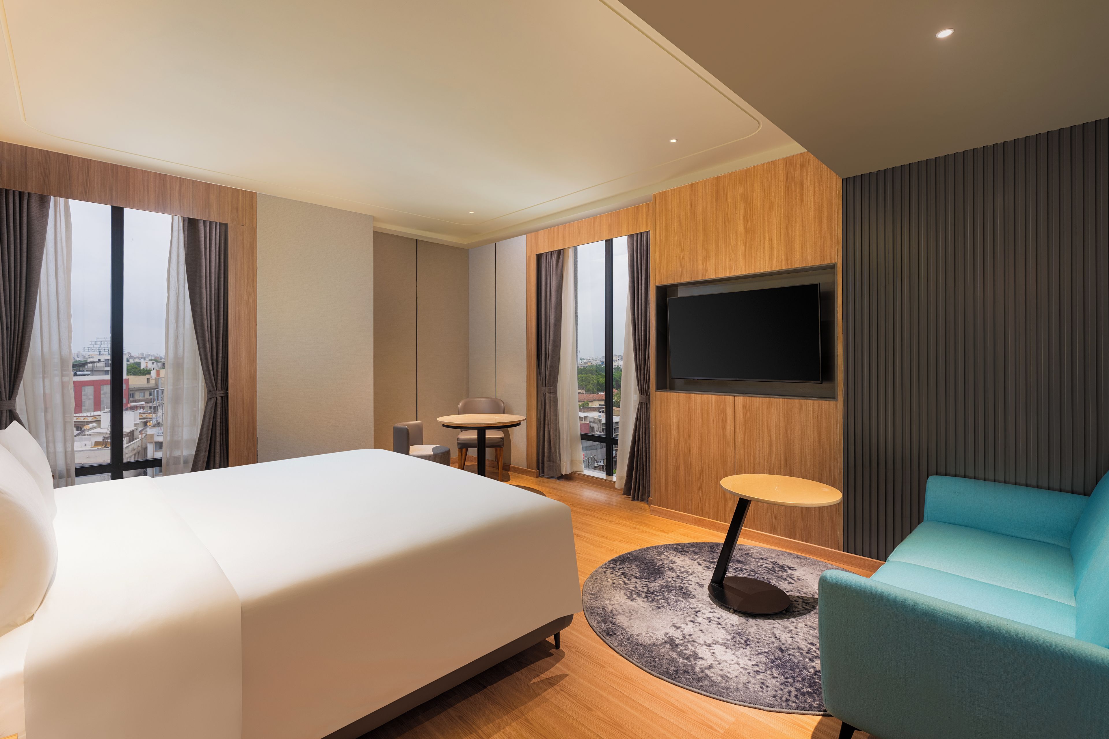 Habitación de hotel con cama, zona de estar con mesa y sillas, televisor de pantalla plana en la pared y ventanas amplias con vistas a la ciudad.