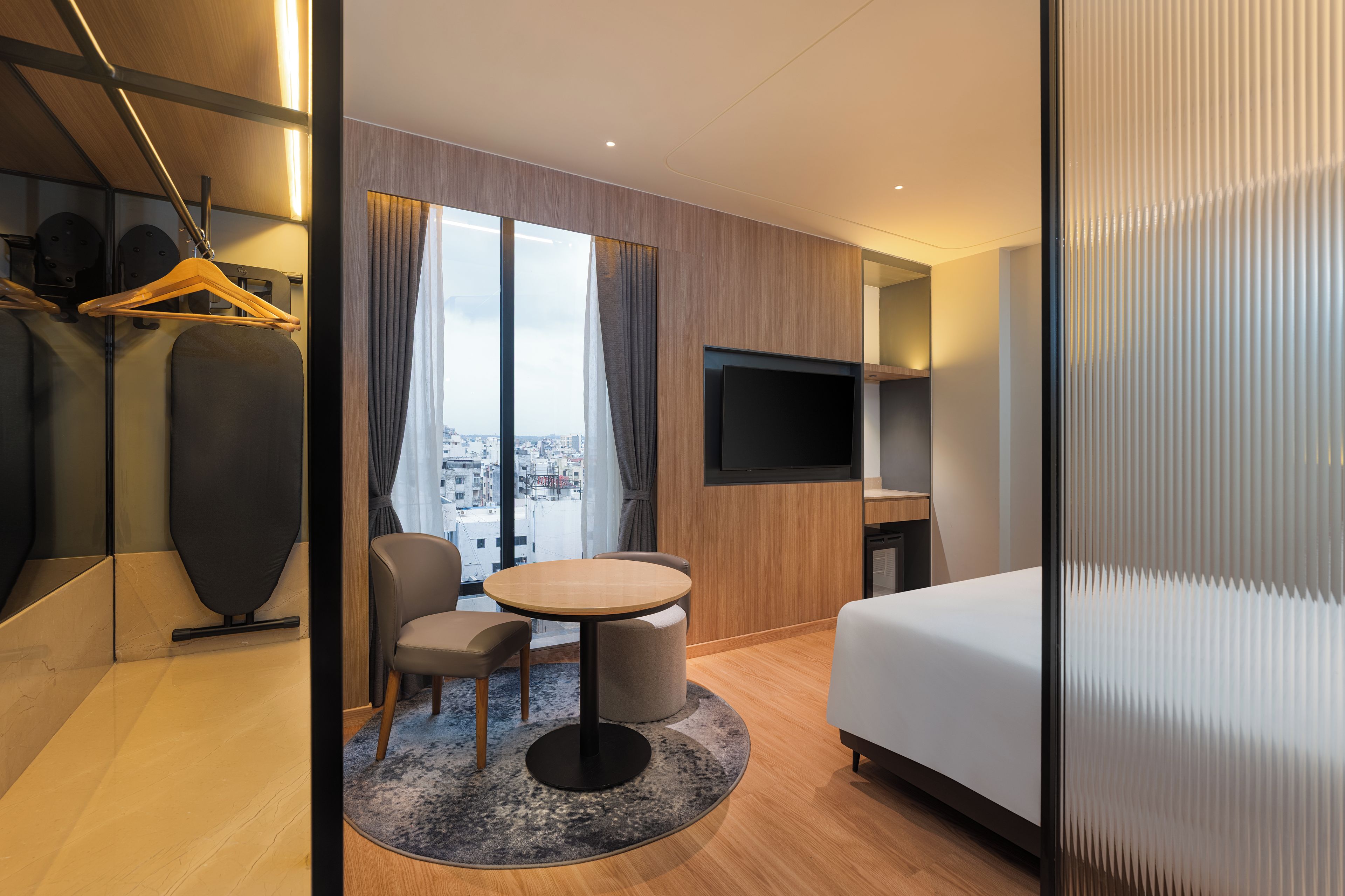 A modern hotel room with a bed, a round table, two chairs, a TV, and a view of the city through large windows.