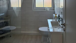 A modern bathroom with a shower, toilet, and sink, featuring a large window and a ceiling-mounted air vent. - Inselruhe - Ihre Auszeit auf Fehmarn (Fehmarn)