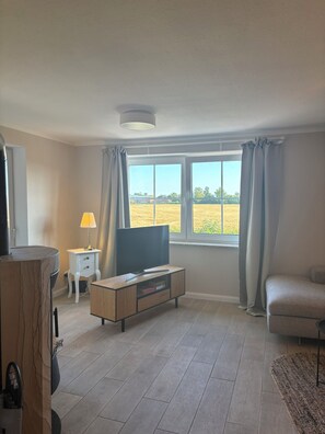 A living room with a television, a sofa, a coffee table, a lamp, and a view of a field through a window. - Inselruhe - Ihre Auszeit auf Fehmarn (Fehmarn)