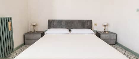 Una cama individual estándar con cabecera gris, dos mesitas de noche con lámparas y un radiador a la izquierda.