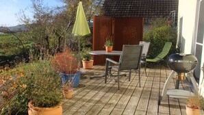 Een terras buiten met een tafel, stoelen, potplanten en een barbecue.