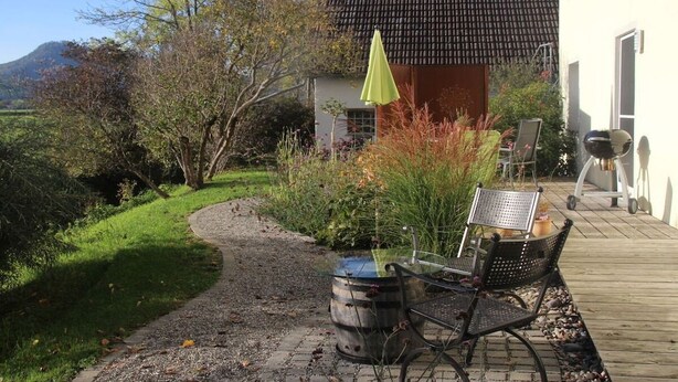 Een terras met een tafel, stoelen, een vat en een klein tuinhuisje met een groene parasol.