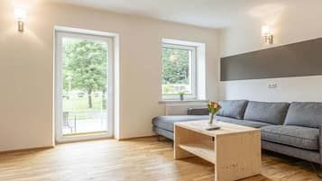 Ein modernes Wohnzimmer mit einem grauen Sofa, einem Holztisch und einem Fenster mit Blick auf Grünflächen.