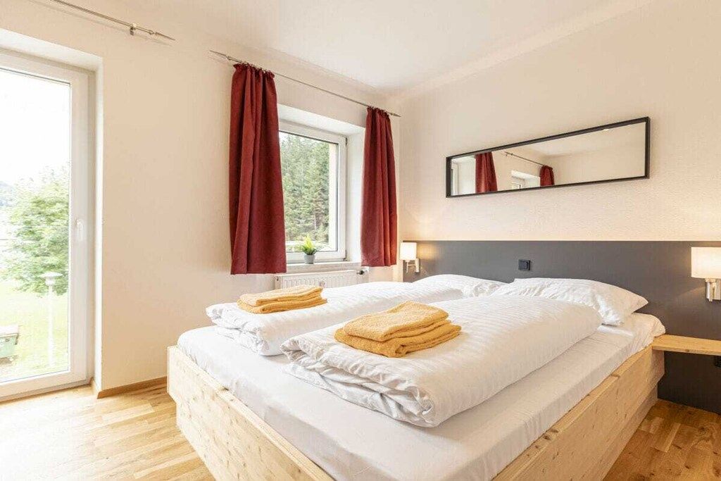 Een tweepersoonsbed met witte lakens en twee opgevouwen handdoeken, een raam met rode gordijnen, een spiegel aan de muur en een nachtkastje met een lamp.