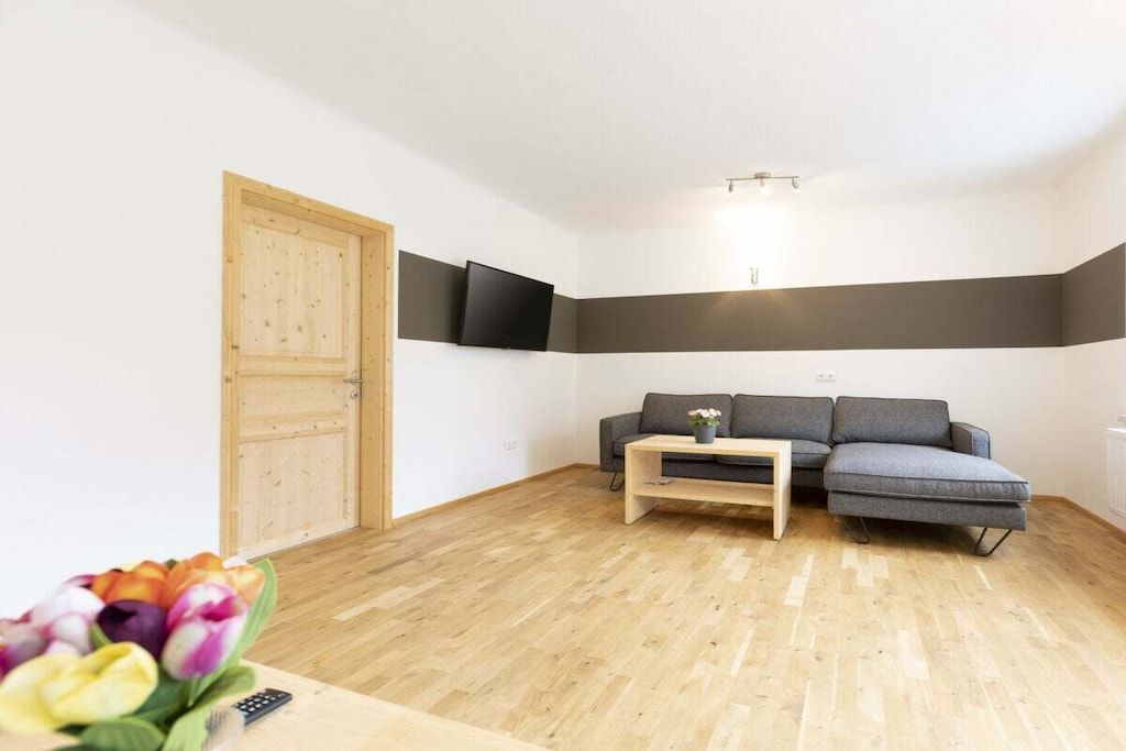 Ein Zimmer mit Holztür, einem grauen Sofa, einem kleinen Tisch und einem an der Wand befestigten Flachbildfernseher.