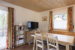 Ein Zimmer mit Holzboden, einem Fernseher auf einem Holzständer, einem Esstisch mit Stühlen, einem Bücherregal und einem Fenster mit Vorhängen.