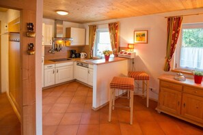 Une cuisine avec des meubles blancs, un îlot central, des tabourets, une machine à café et une fenêtre avec des rideaux.