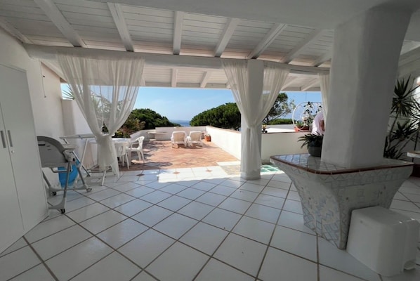 Un patio techado con piso de baldosas, mobiliario blanco y vista al océano.