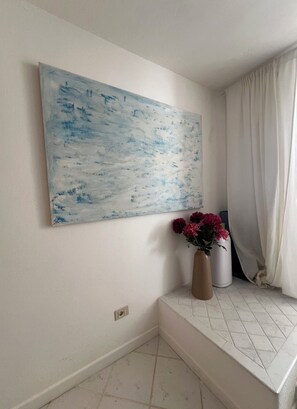 Ein Zimmer mit einem großen abstrakten Gemälde an der Wand, einer Vase mit Blumen auf einem Fensterbrett und einem weißem, halb zurückgezogenen Vorhang.