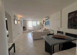 A modern living room with a dining table, sofa, and a large window. - views and big terrace  steps from the beach (Baja Sardinia)